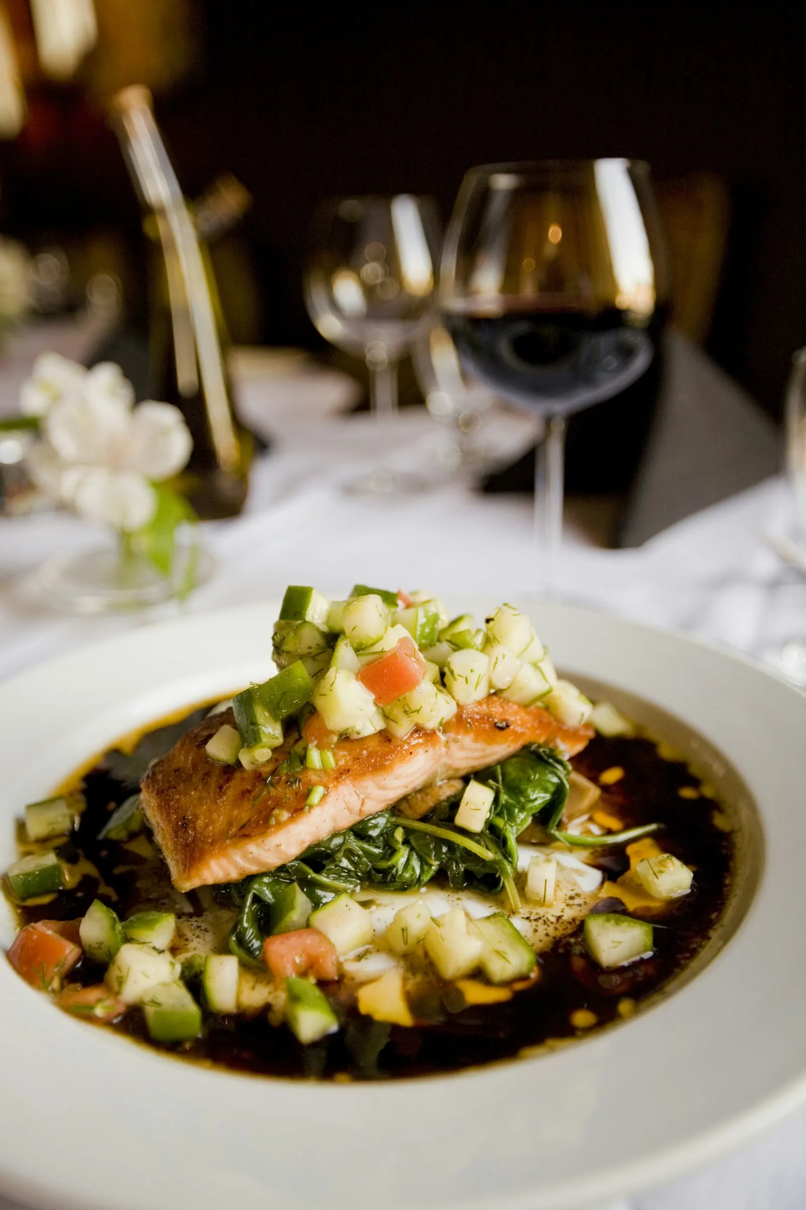 Angerichteter Fischgang mit Gemüsewürfeln und dunklem Jus, im Hintergrund Weingläser